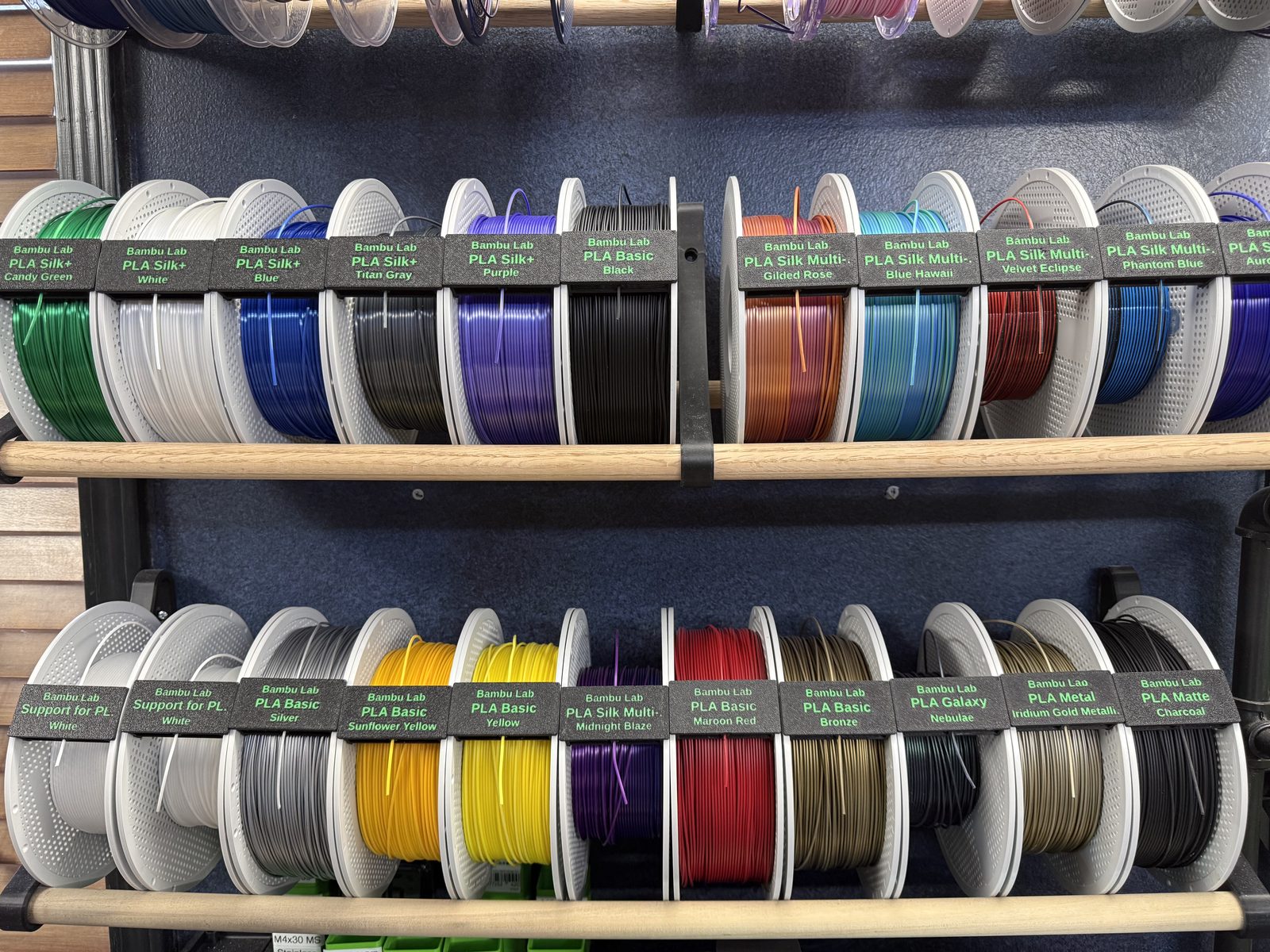 Rows of filament spools on a shelf, each with a readable label clip showing brand, type and color